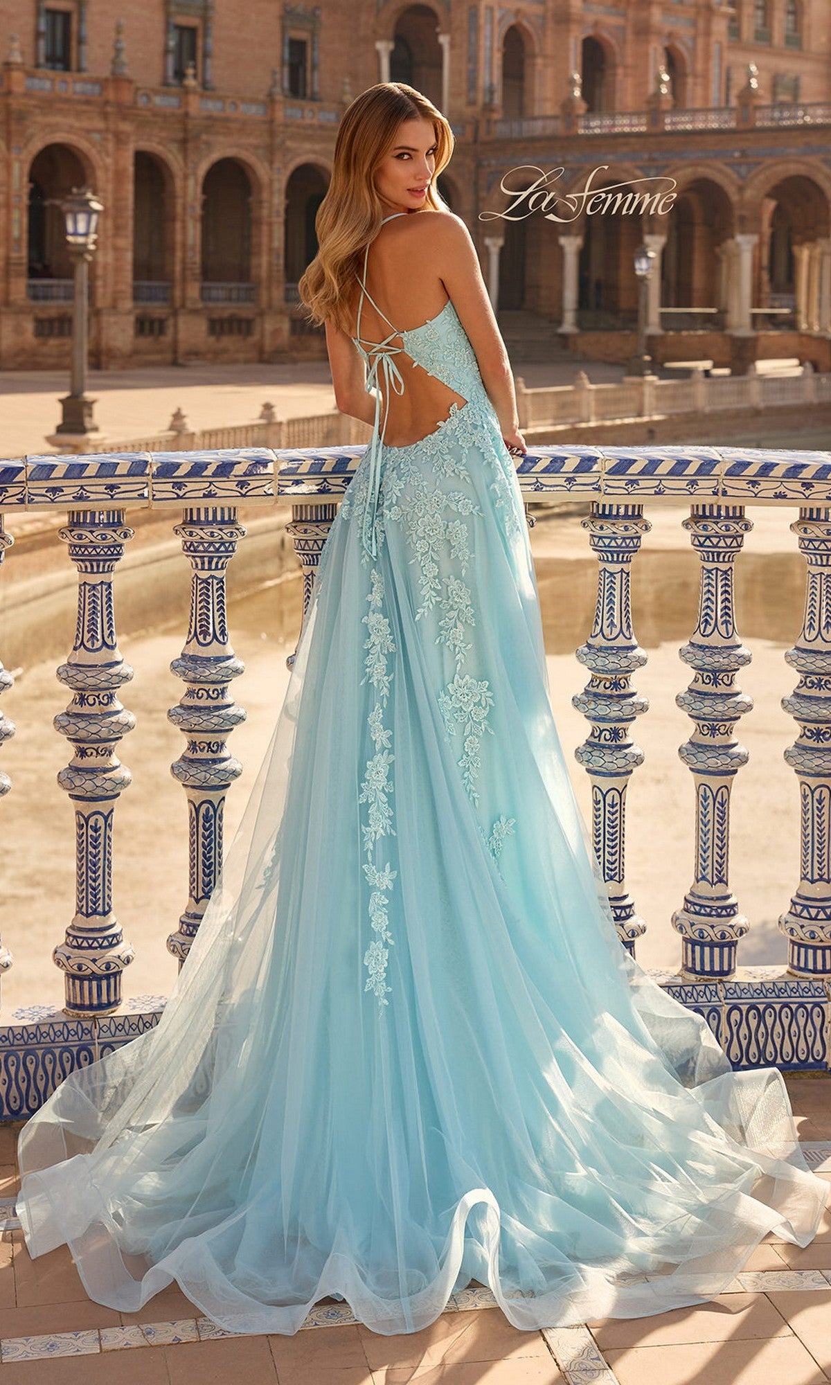 Wearing the La Femme 33715 long prom dress in light blue tulle with lace embroidery, a woman stands by a decorative railing in an outdoor plaza.